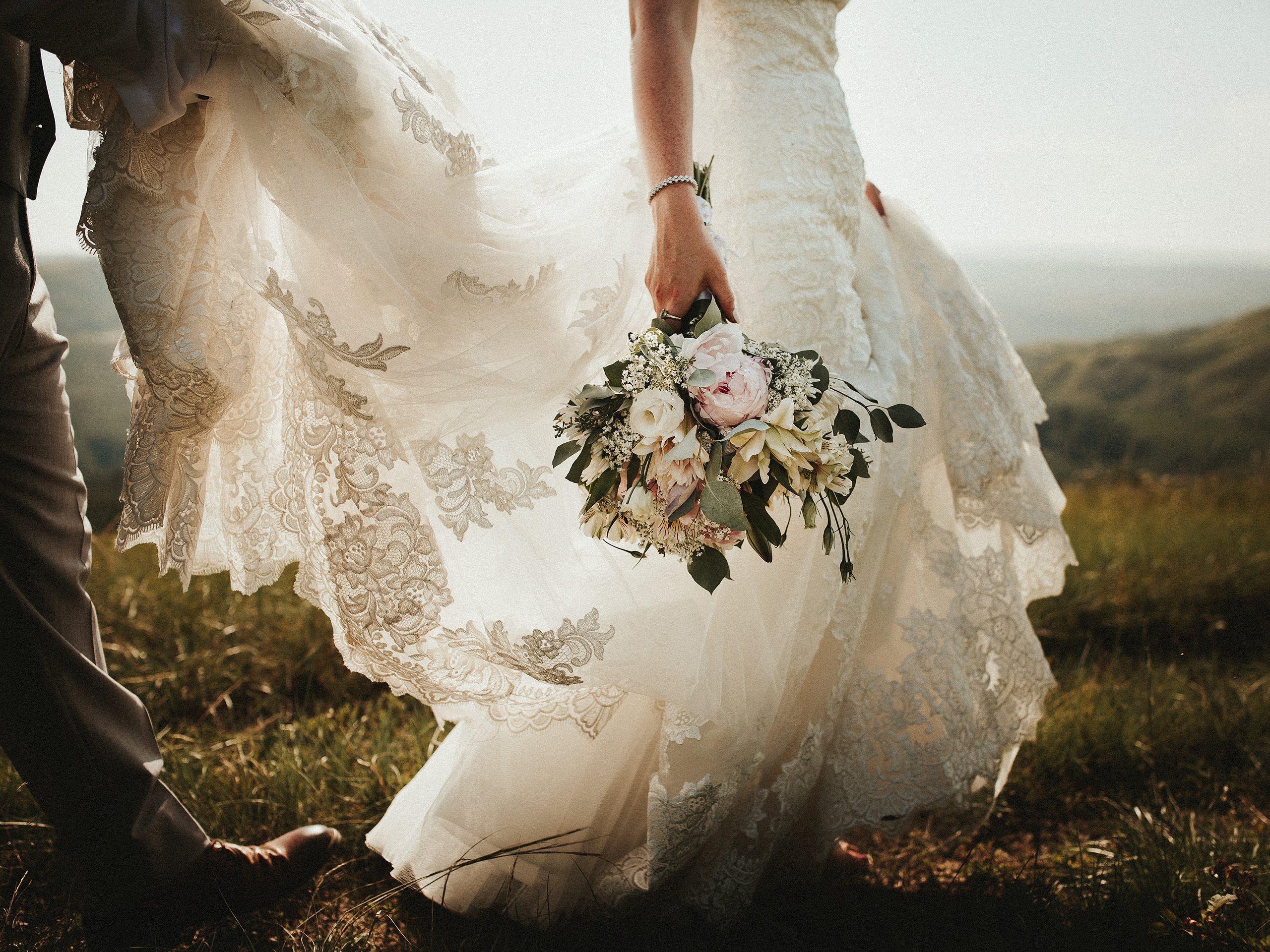 Groom holds brides dress as they walk up a mountain.